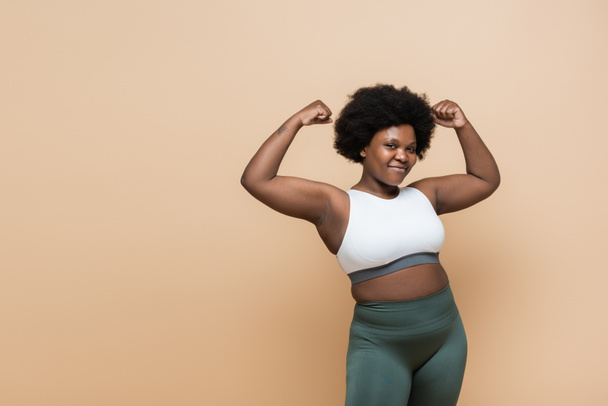 cheerful african american plus size woman in crop top showing power gesture isolated on beige - Photo, image