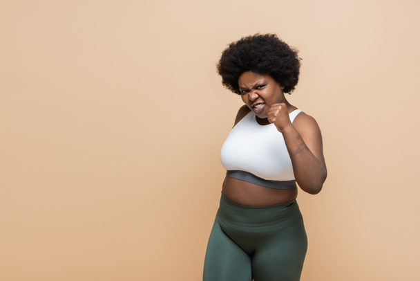 emotional african american plus size woman in crop top showing clenched fist isolated on beige - Foto, imagen
