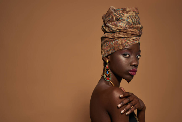 Side view of serious elegant black girl wearing traditional african outfit and accessories looking at camera. Cropped of beautiful young slim woman. Isolated on orange background in studio. Copy space - Fotó, kép