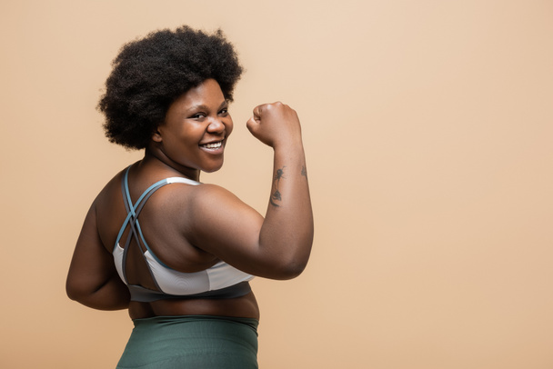 татуйована афроамериканка плюс розмір жінка у верхній частині врожаю посміхається і показує силовий жест ізольовано на бежевому
 - Фото, зображення