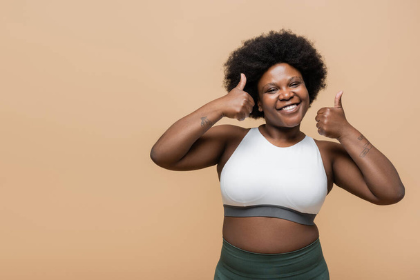 Χαρούμενη Αφροαμερικανή συν μέγεθος γυναίκα στην κορυφή των καλλιεργειών δείχνει τους αντίχειρες επάνω απομονωμένη σε μπεζ - Φωτογραφία, εικόνα