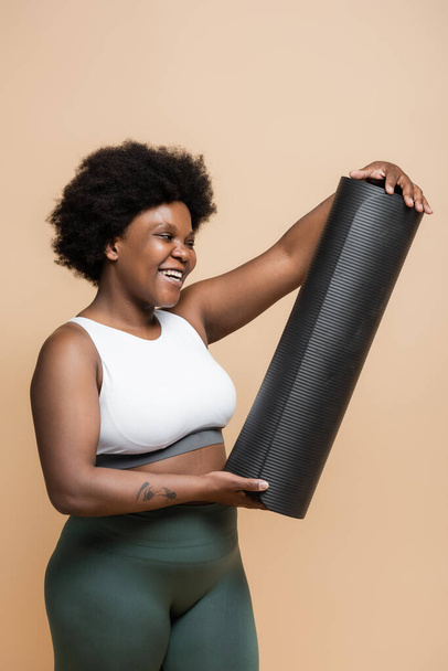 happy african american plus size woman in crop top looking at fitness mat isolated on beige - Foto, afbeelding