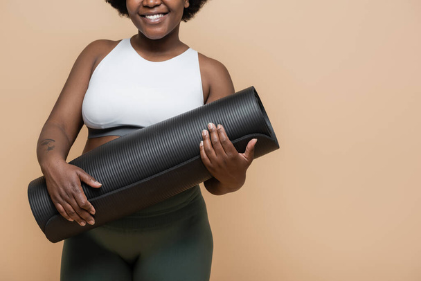 cropped view of happy african american plus size woman with tattoo holding fitness mat isolated on beige - Foto, Imagem