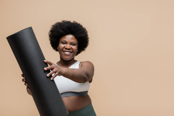 χαμογελαστή και σγουρή αφρικανή γυναίκα συν μέγεθος στην καλλιέργεια κορυφή εκμετάλλευση στρώμα γυμναστικής που απομονώνονται σε μπεζ - Φωτογραφία, εικόνα