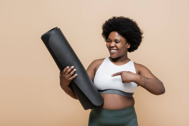 happy african american plus size woman in crop top pointing at fitness mat isolated on beige - Foto, immagini