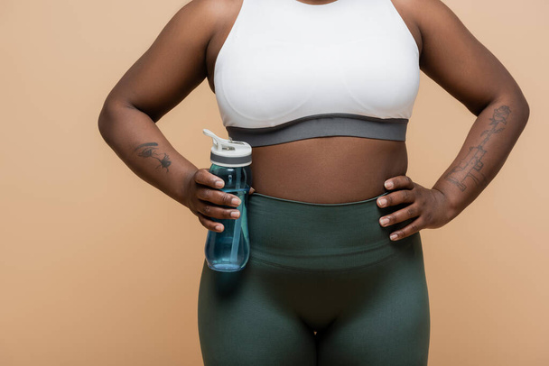 cropped view of tattooed african american plus size woman in crop top holding sports bottle with water isolated on beige - Foto, Imagen