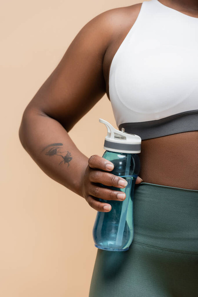 обрізаний вид татуйованої афроамериканки плюс розмір жінки, що тримає спортивну пляшку ізольовано на бежевому
 - Фото, зображення
