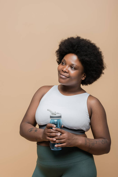 dreamy african american plus size woman in crop top holding sports bottle with water isolated on beige - Foto, Imagen