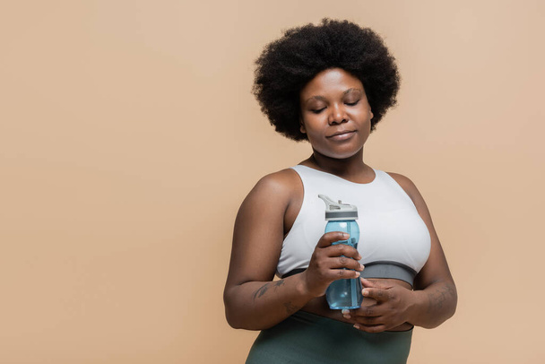 кучерява афроамериканка плюс розмір жінка у верхній частині врожаю тримає спортивну пляшку з водою ізольовано на бежевому
 - Фото, зображення