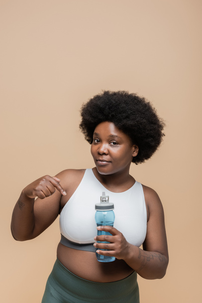 african american plus size woman in crop top pointing at sports bottle with water isolated on beige - Foto, immagini