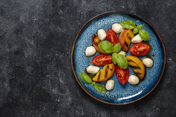 Grilled peach salad with mozzarella, tomato, green basil and sauce. Fresh gourmet salad in plate on black background, close up, top view, copy space - Photo, Image