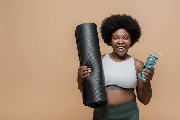 positive african american plus size woman in crop top holding fitness mat and sports bottle isolated on beige - Zdjęcie, obraz