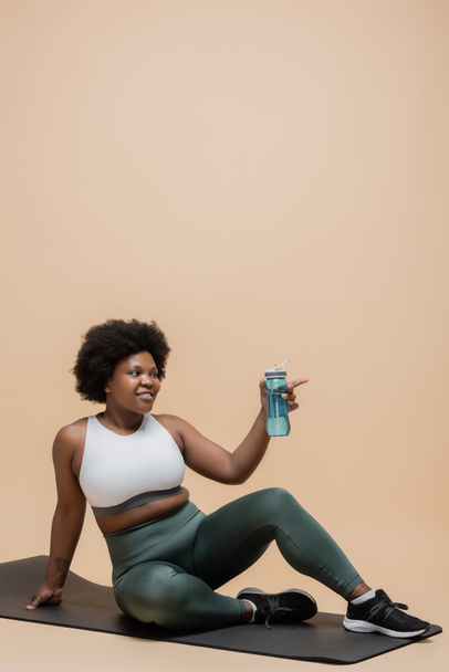 positive african american plus size woman in sportswear sitting on fitness mat and holding sports bottle on beige - Fotó, kép