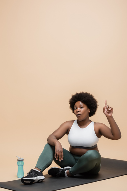 кудрявый африканский американец плюс размер женщины в спортивной одежде сидя на фитнес-коврик возле спортивной бутылки и указывая на бежевый - Фото, изображение