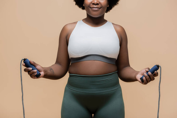 cropped view of cheerful african american plus size woman in sportswear holding skipping rope isolated on beige  - Zdjęcie, obraz