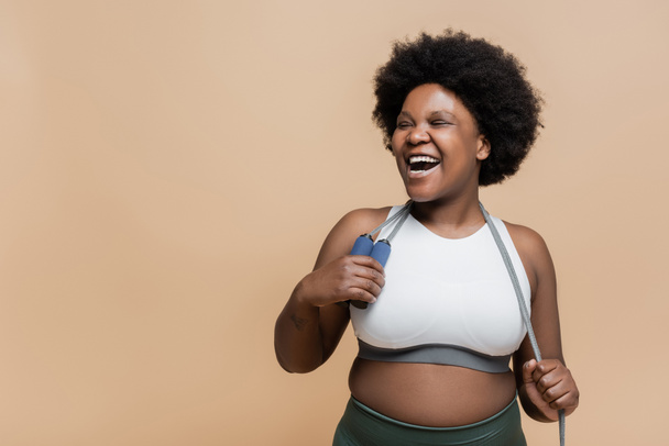 excited african american plus size woman in sportswear holding skipping rope isolated on beige  - Foto, Imagen