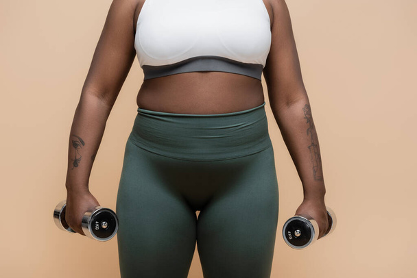 обрезанный вид на татуированный африканский американец плюс размер женщины в спортивной одежде держа гантели изолированы на бежевый  - Фото, изображение