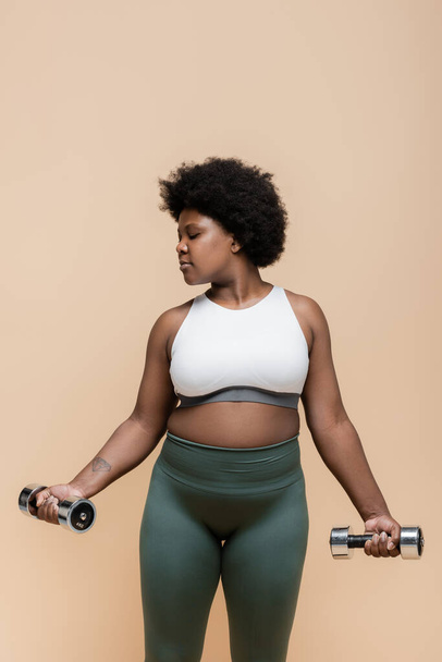 татуированные африканские американец плюс размер женщины в спортивной форме упражнения с гантели изолированы на бежевый  - Фото, изображение