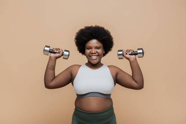 happy african american plus size woman in sportswear exercising with dumbbells isolated on beige  - Fotografie, Obrázek