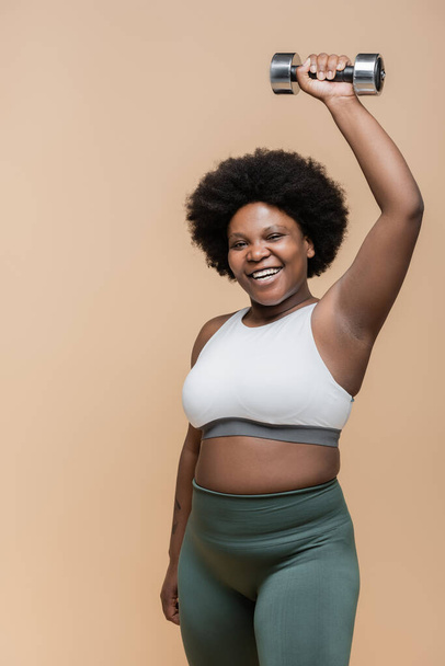 счастливый африканский американец плюс размер женщины в спортивной форме упражнения с гантели изолированы на бежевый  - Фото, изображение
