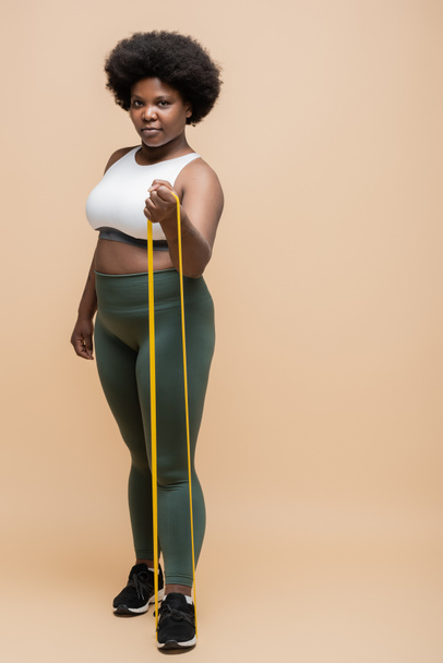 full length of african american plus size woman in sportwear exercising with resistance band on beige - Foto, Bild