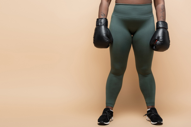 καλλιεργημένη άποψη της αφρικανικής Αμερικής συν μέγεθος γυναίκα με γάντια πυγμαχίας στέκεται σε κολάν σε μπεζ - Φωτογραφία, εικόνα
