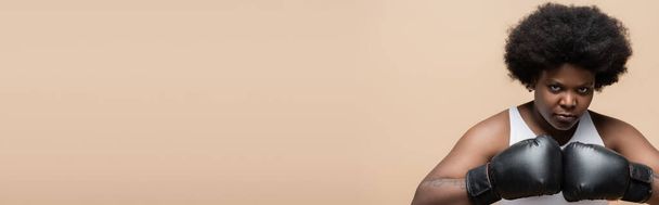 ισχυρή αφρικανή αμερικανίδα συν μέγεθος γυναίκα με γάντια πυγμαχίας απομονώνονται σε μπεζ, banner - Φωτογραφία, εικόνα