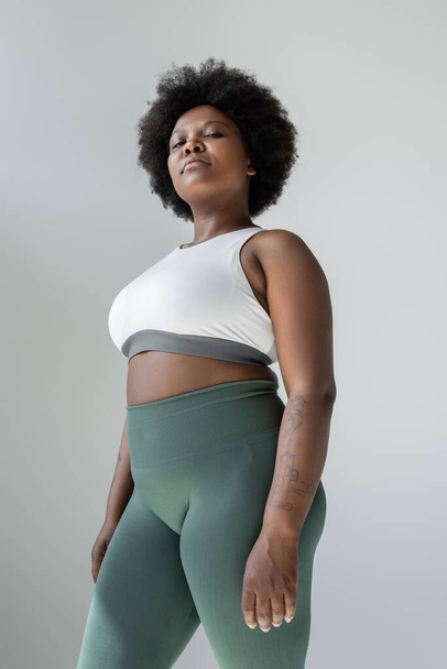 low angle view of tattooed african american plus size woman in sportswear standing isolated on grey - Foto, afbeelding