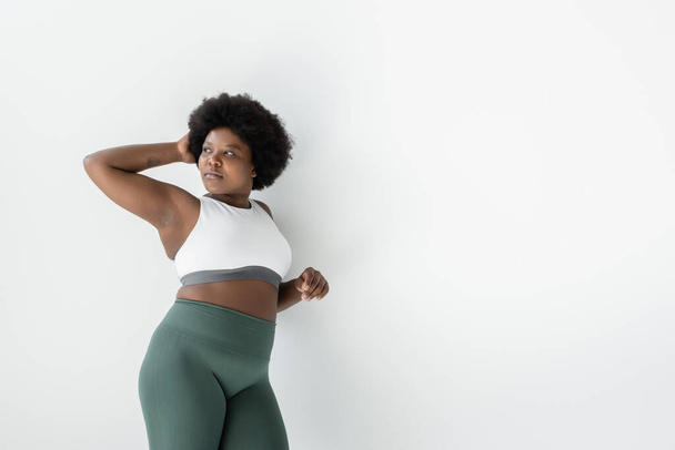 curly african american plus size woman in sportswear standing near white wall  - Foto, afbeelding