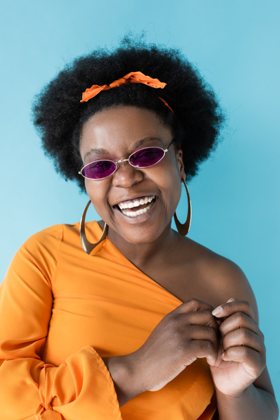 positive african american plus size woman in sunglasses and hoop earrings isolated on blue - Foto, Imagem
