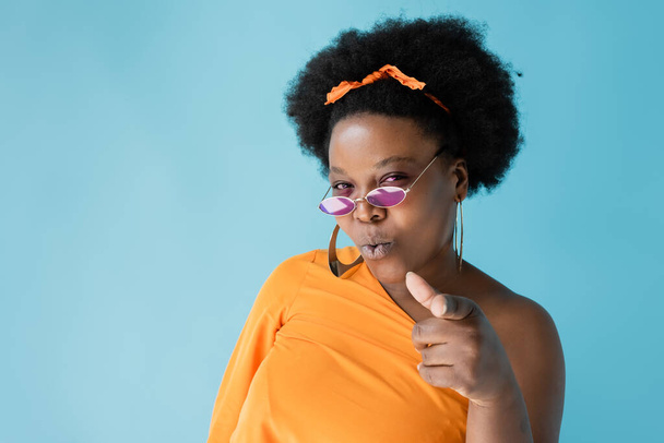 curly african american woman in pink sunglasses pointing with finger isolated on blue - Valokuva, kuva