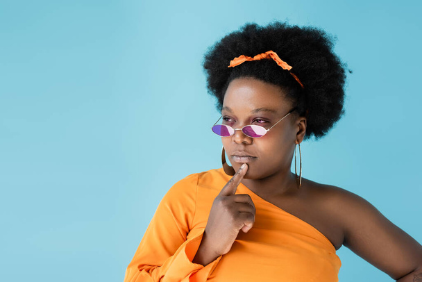 pensive african american body positive woman in pink sunglasses isolated on blue - Foto, Bild