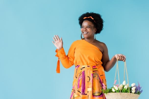cheerful african american body positive woman holding straw bag with flowers isolated on blue - Foto, Bild