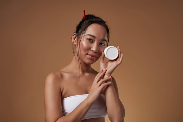 Smiling stylish asian girl holding container with cosmetic cream and looking at camera. Attractive young brunette woman wear tank top. Body and skin care. Orange background. Studio shoot. Copy space - Photo, Image