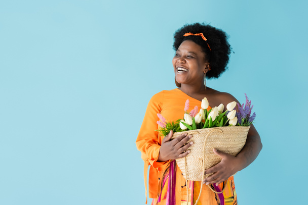позитивна афроамериканка тримає солом'яний мішок з квітами ізольовано на синьому
 - Фото, зображення
