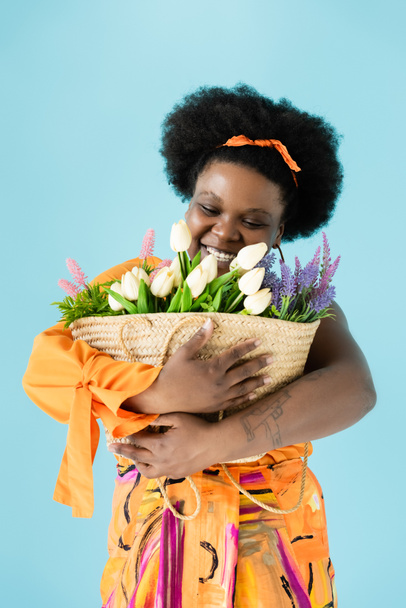 happy african american body positive woman hugging straw bag with flowers isolated on blue - Photo, image