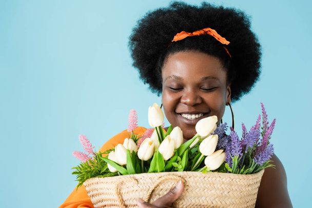 Χαρούμενη Αφροαμερικανή θετική γυναίκα που αγκαλιάζει αχυρένια τσάντα με λουλούδια απομονωμένα στο μπλε - Φωτογραφία, εικόνα