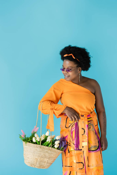 счастливая африканская американское тело позитивная женщина в солнцезащитных очках держа соломенный мешок с цветами изолированы на синий - Фото, изображение
