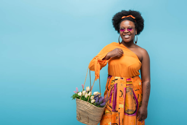 cheerful african american body positive woman in sunglasses holding straw bag with flowers isolated on blue - Foto, Bild