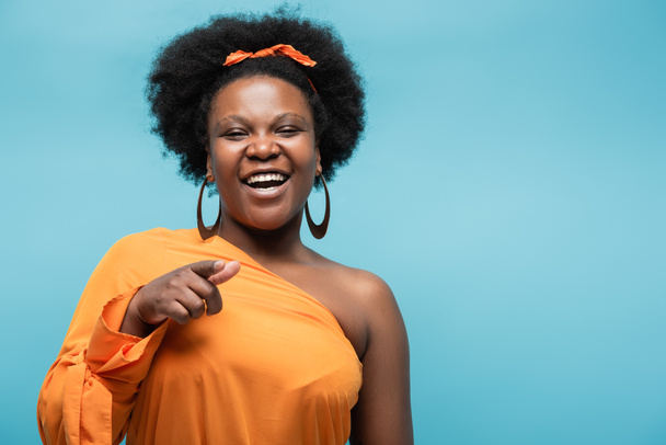 χαμογελαστή αφροαμερικανίδα θετική στο σώμα γυναίκα με πορτοκαλί φόρεμα και σκουλαρίκια στεφάνης που δείχνουν με το δάχτυλο απομονωμένο στο μπλε - Φωτογραφία, εικόνα