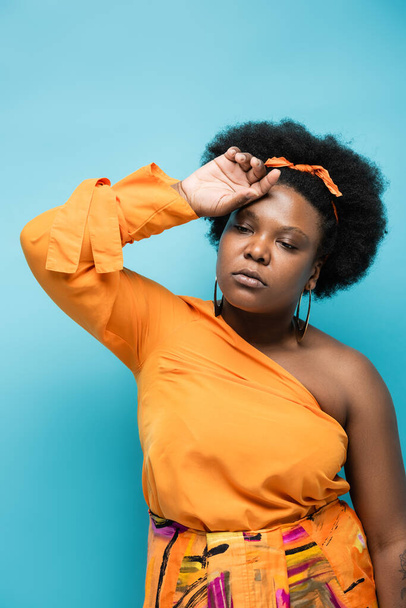 curly african american body positive woman in orange dress and hoop earrings feeling unwell isolated on blue - Foto, afbeelding