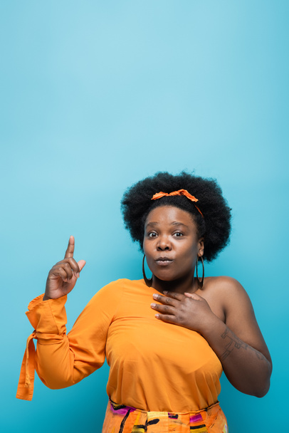 tattooed african american body positive woman in orange dress and hoop earrings pointing with finger isolated on blue - Foto, afbeelding