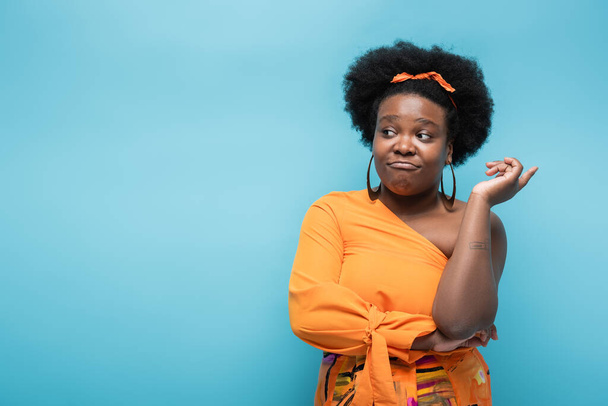 confused african american body positive woman in orange dress and hoop earrings isolated on blue - Fotó, kép