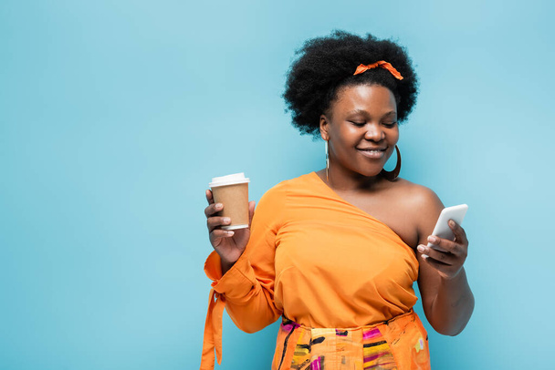 pleased african american body positive woman in hoop earrings holding paper cup and using smartphone isolated on blue - 写真・画像