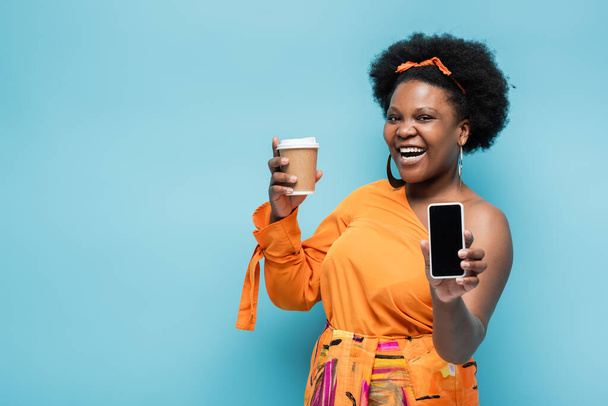 happy african american body positive woman holding paper cup and smartphone with blank screen isolated on blue - Fotoğraf, Görsel