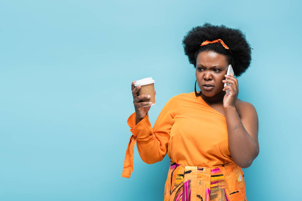 worried african american body positive woman holding paper cup and and talking on smartphone isolated on blue - Fotografie, Obrázek