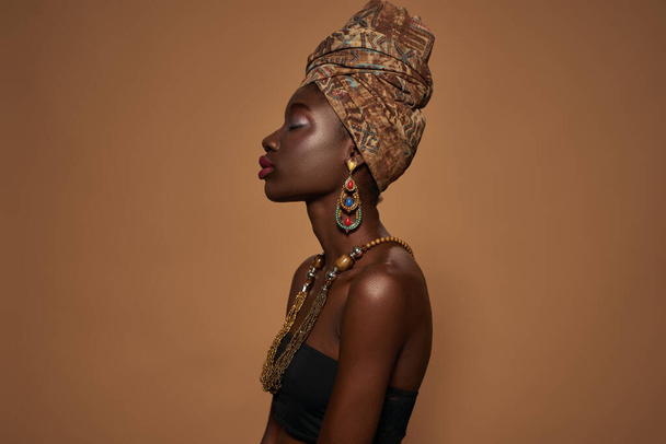 Side view of dreamy stylish black girl with closed eyes wearing traditional african outfit and accessories. Attractive young slim woman. Isolated on orange background. Studio shoot. Copy space - Foto, Imagem
