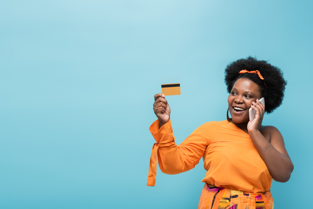 Χαρούμενη Αφροαμερικανή θετική γυναίκα με σκουλαρίκια στεφάνης κρατώντας πιστωτική κάρτα και μιλώντας σε smartphone απομονωμένο στο μπλε - Φωτογραφία, εικόνα