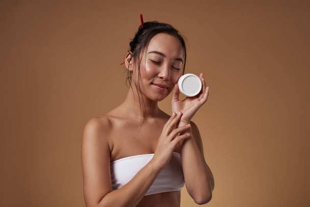 Fashionable asian girl with closed eyes holding container with cosmetic cream. Pretty young brunette slim woman wear tank top. Body and skin care. Isolated on orange background in studio. Copy space - Foto, afbeelding