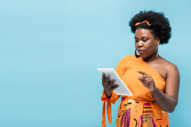 curly african american body positive woman in hoop earrings holding digital tablet isolated on blue - Foto, afbeelding